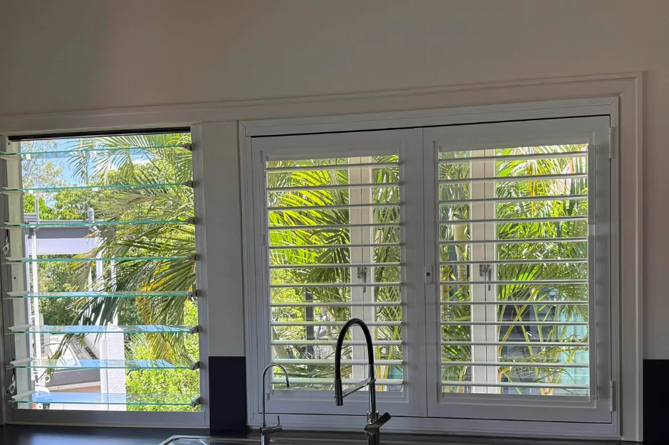 A kitchen window with white Guardian shutters shut, the key locks showing and the blades tilted open to the palms outside