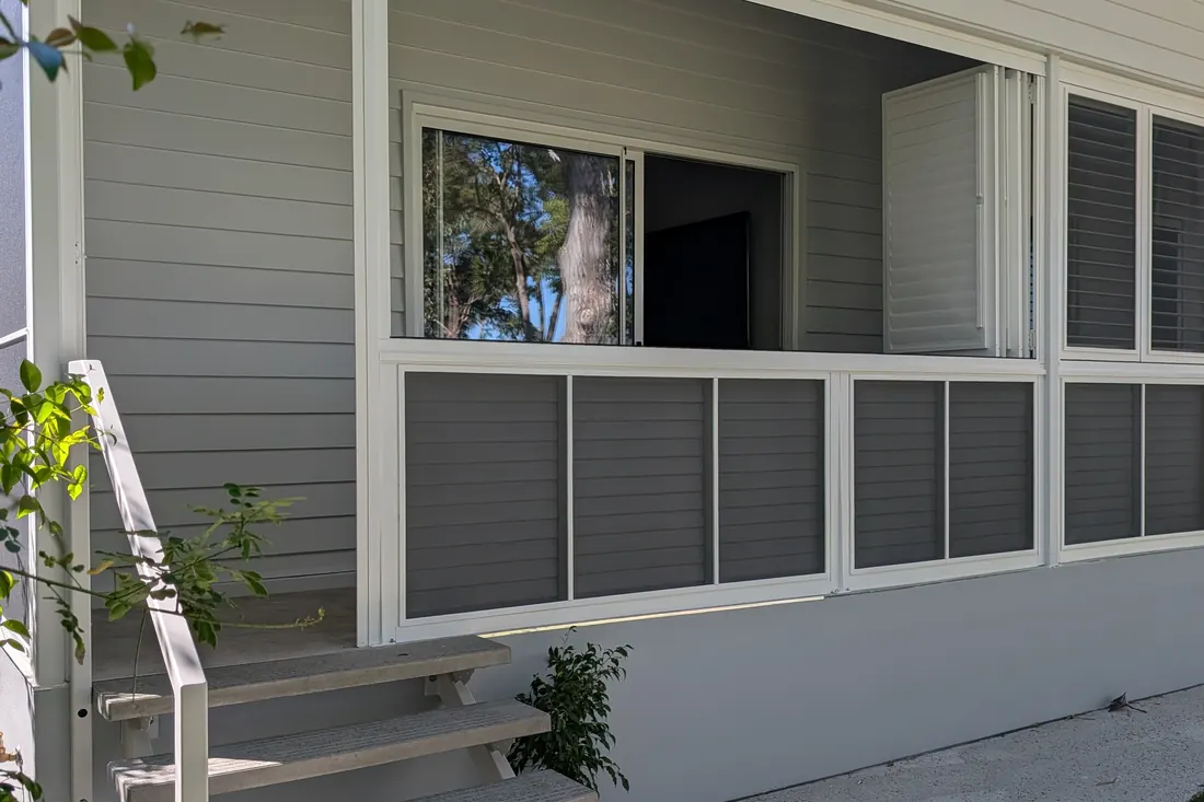 A veranda closed in with grey Guardian shutters, one upper panel swung open beside the window